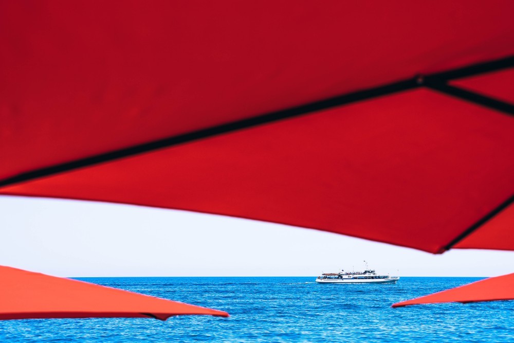 Boat floating in the beautiful blue sea, as seen behind red beach umbrellas.