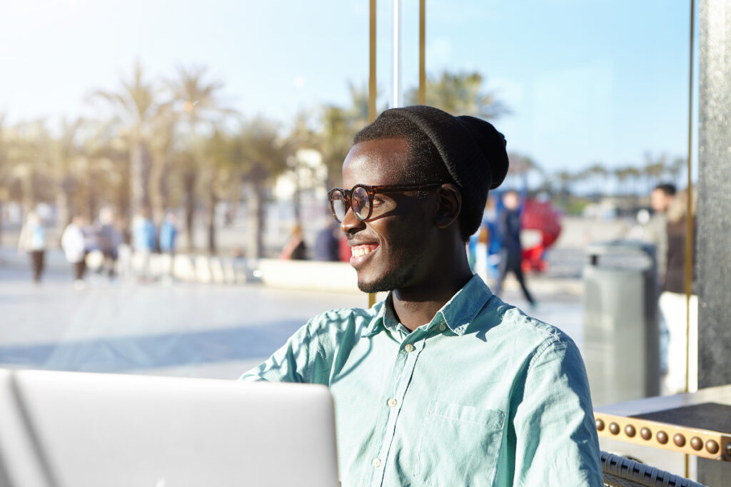 relaxed carefree dark skinned student dressed in fashionable clothing using free high speed internet connection on generic laptop pc while sitting at light spacious cafe waiting for his lunch